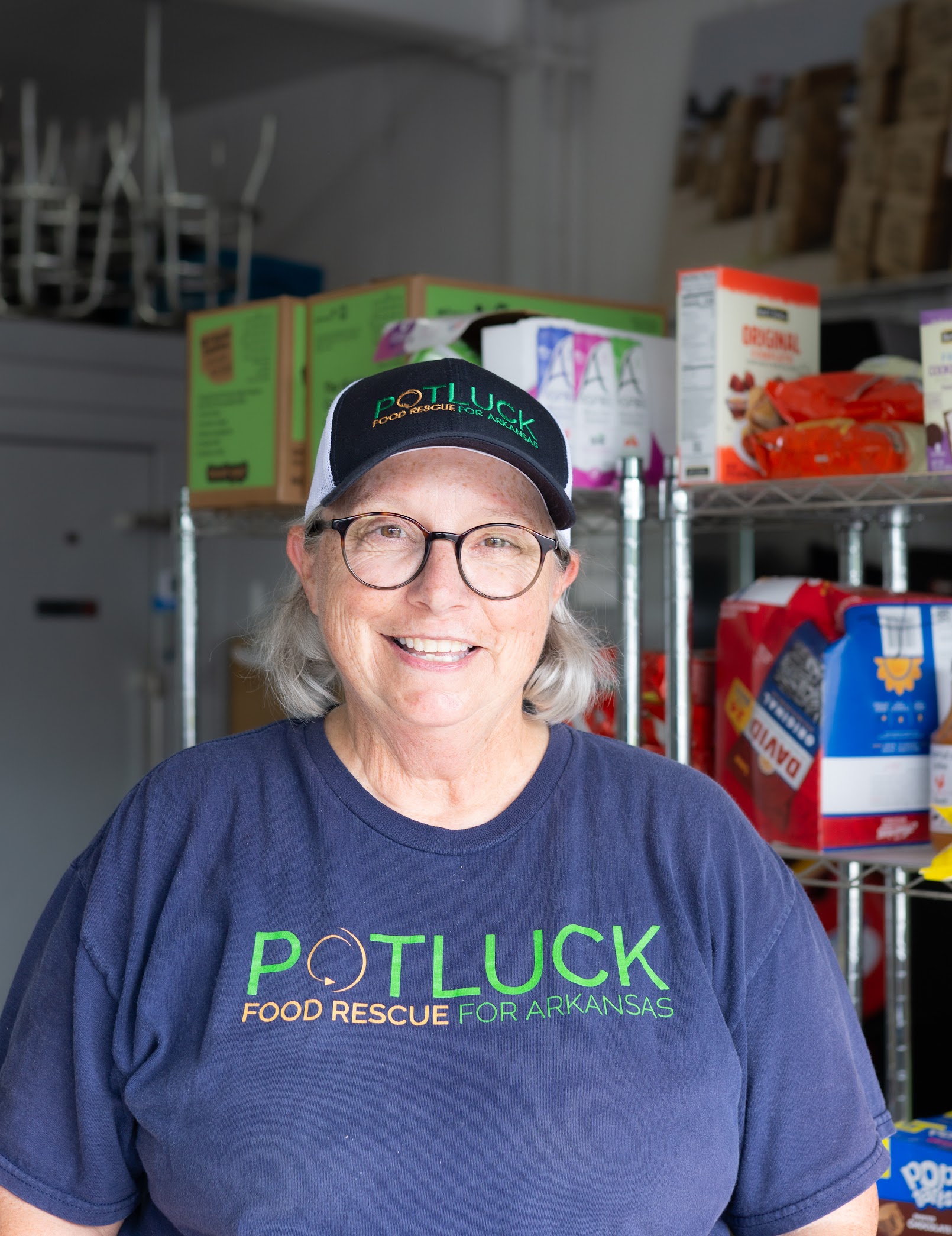 DSC09847 Smiling woman wearing glasses, a POTLUCK Food Rescue for Arkansas shirt, and a matching hat stands in front of shelves filled with food items and boxes in what appears to be a food pantry.