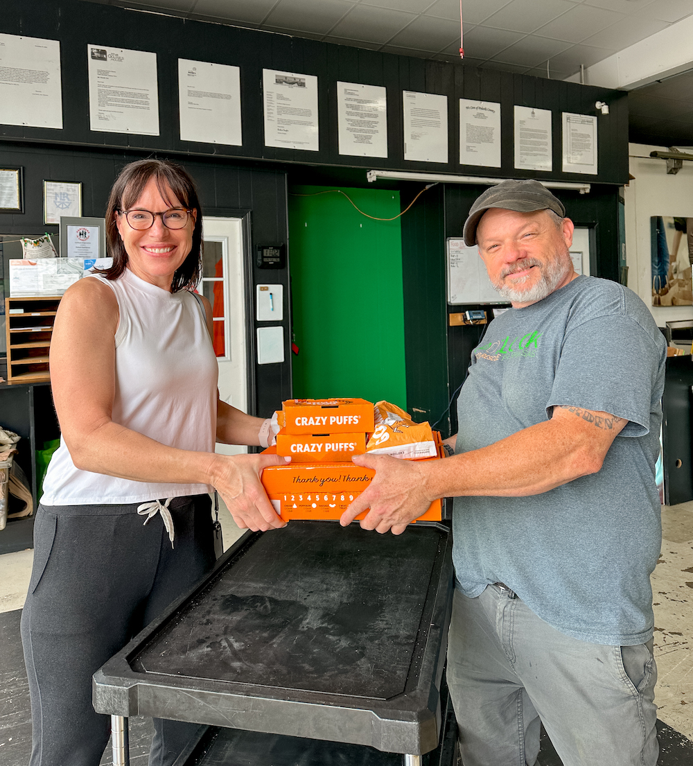Howcanyouhelp Two people smile while holding orange boxes labeled Crazy Puffs in an indoor setting. The woman wears glasses and a sleeveless top; the man has a gray beard, T-shirt, and cap. They stand by a black cart.