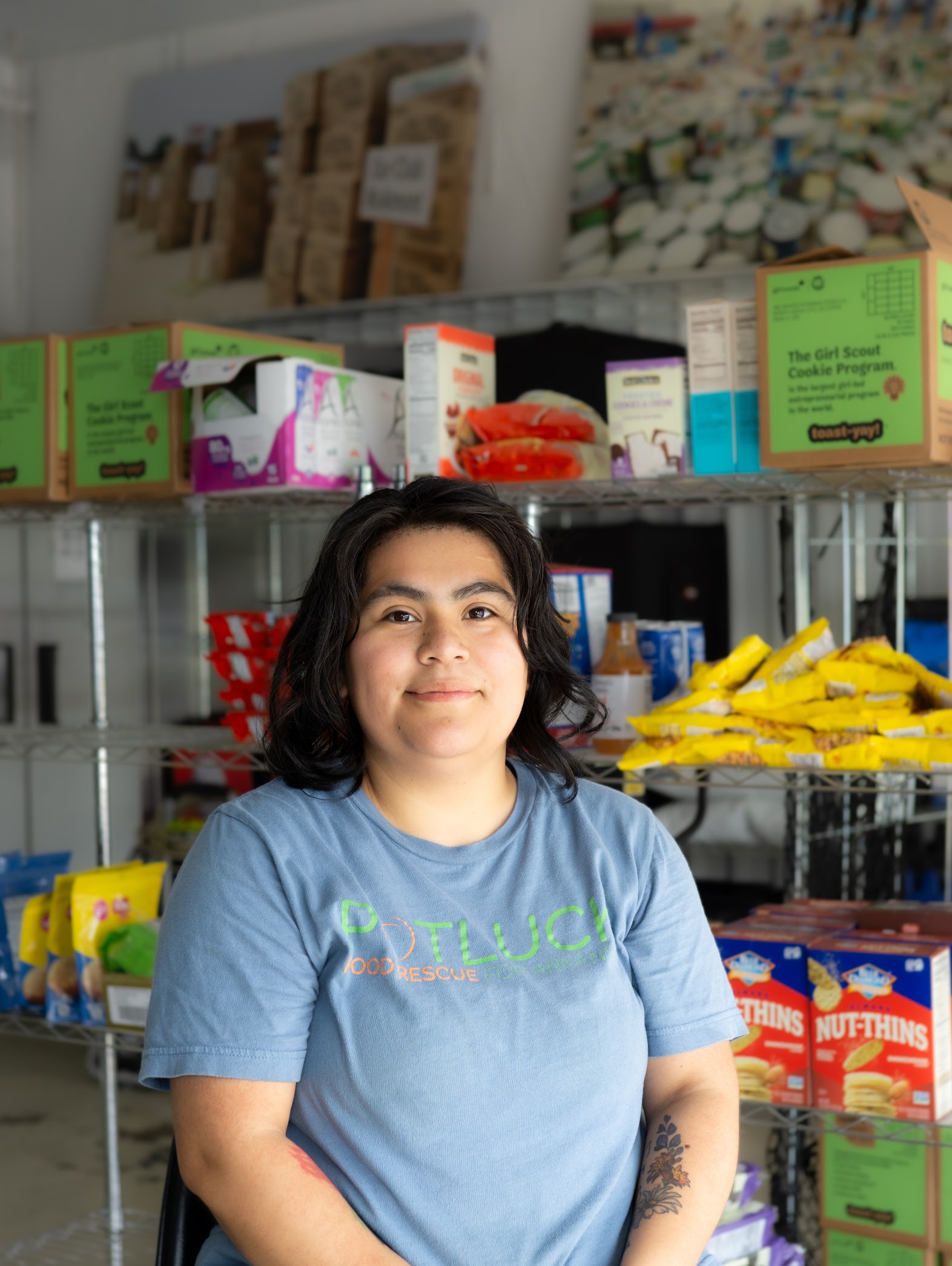 Jackyheadshot1 A woman with dark hair and a blue “Potluck Food Rescue” shirt sits smiling in front of shelves stocked with various food items and cardboard boxes in a food pantry or storage area.