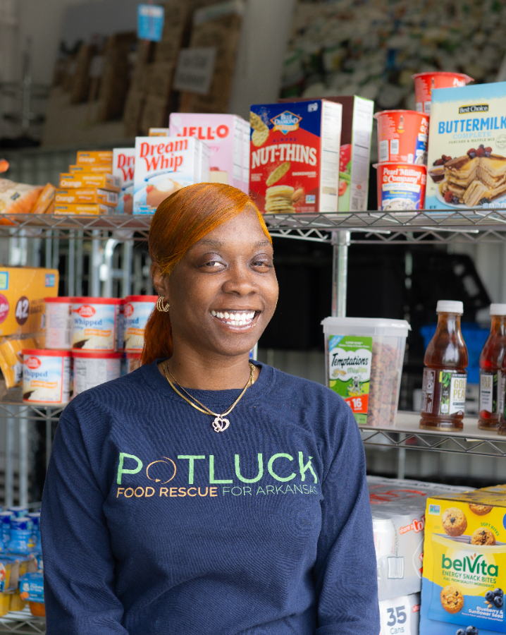 Myaheadshot (1) A woman with orange hair and a navy Potluck Food Rescue for Arkansas shirt smiles in front of shelves stocked with various boxed and canned food items.