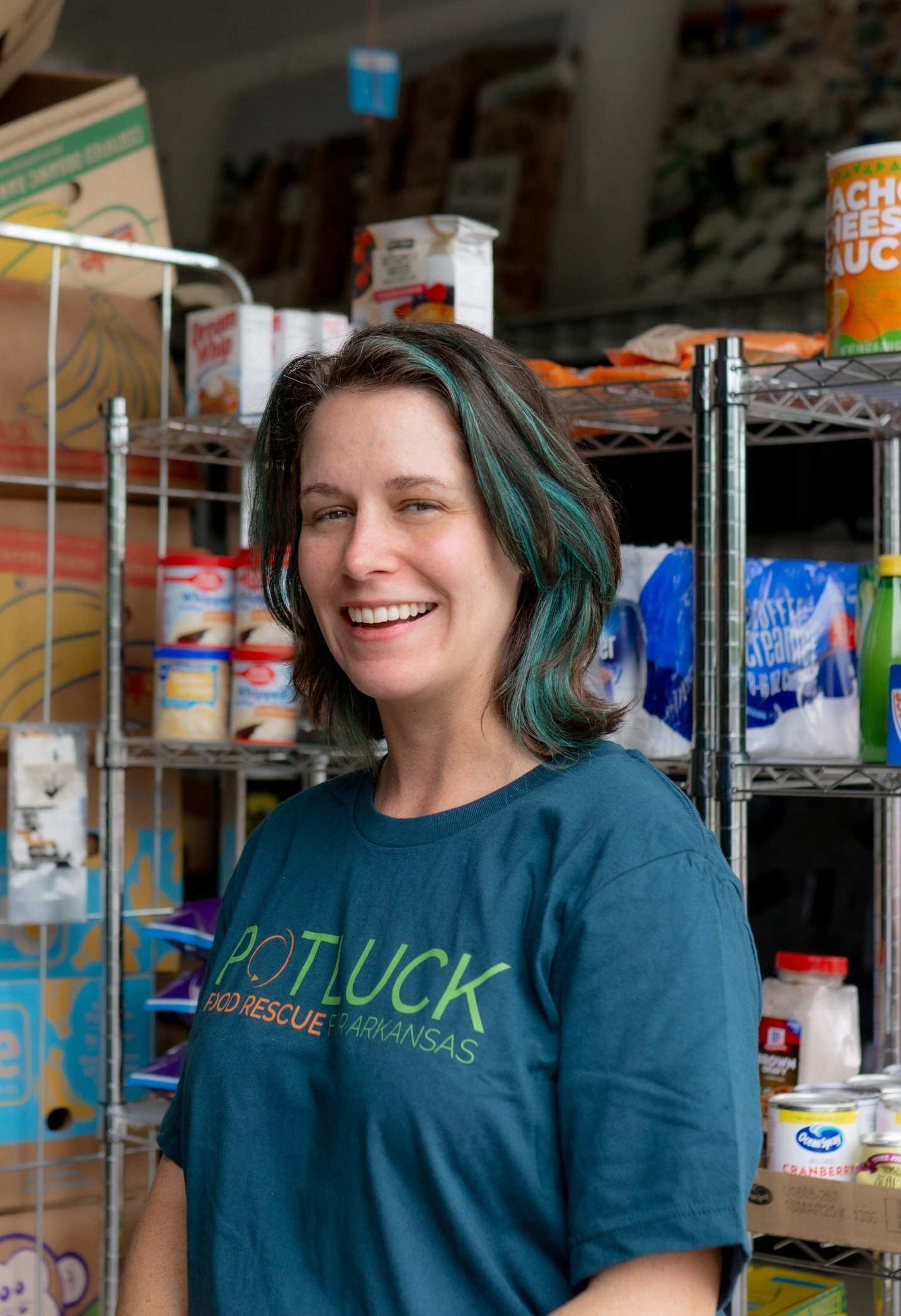 Potluck Marianne A woman with shoulder-length hair wearing a POTLUCK FOOD RESCUE ARKANSAS shirt smiles while standing in front of metal shelves filled with food items and supplies.