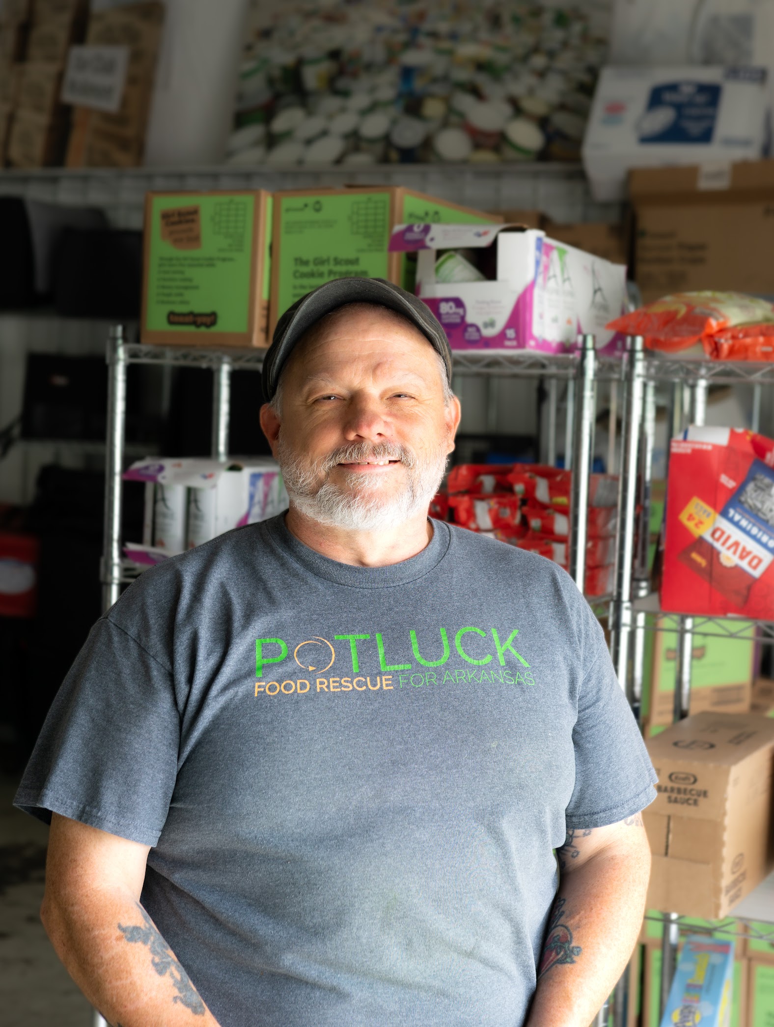 Robertheadshot2 A smiling man with a gray beard and mustache stands in front of metal shelves filled with boxes. He wears a gray POTLUCK FOOD RESCUE FOR ARKANSAS t-shirt and a cap. The background shows more boxes and supplies.