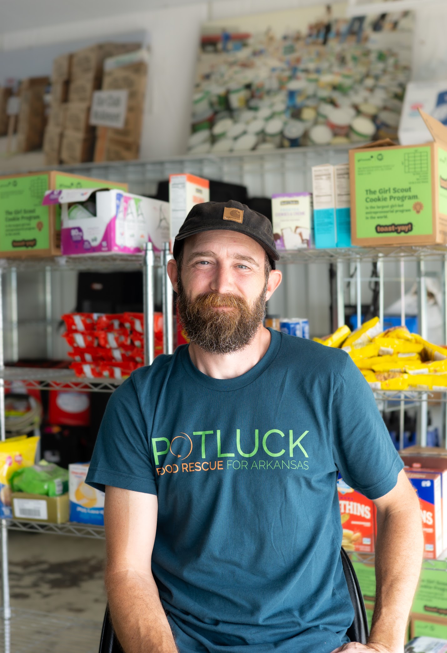 Seanheadshot1 A bearded man in a “Potluck Food Rescue for Arkansas” shirt sits and smiles in front of shelves stocked with assorted food boxes and supplies.