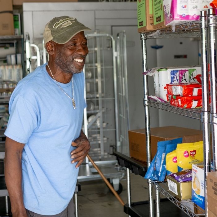 Teamwork Plates Landfills A man in a light blue shirt and tan cap stands and smiles next to metal shelves filled with various packaged goods in a storage or supply room.