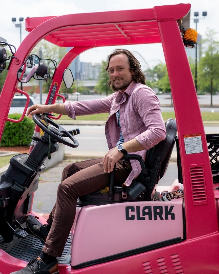 Teamwork Potluck (2) A man wearing a pink shirt and brown pants sits and smiles while driving a pink forklift labeled CLARK in an outdoor parking lot area.