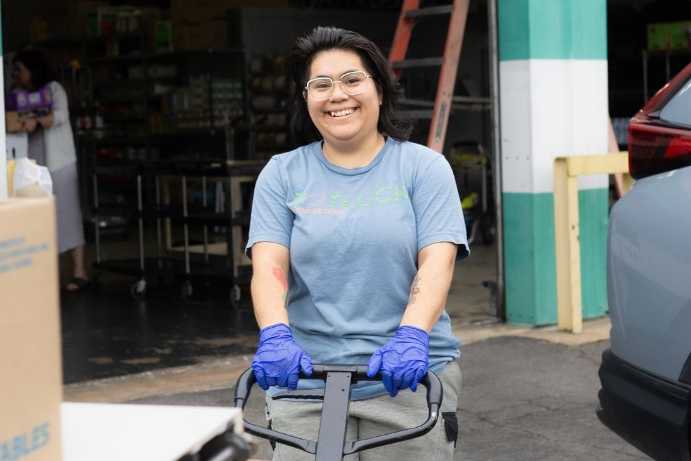 Teamwork Potluck (3) A smiling person wearing glasses, a blue shirt, and blue gloves pushes a cart outside a building, with boxes nearby and another person blurred in the background.