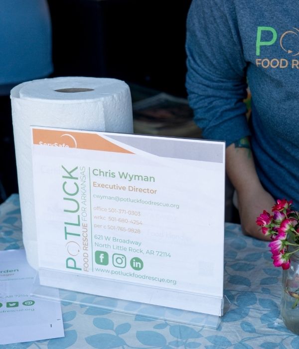 Teamwork Potluck (4) A sign for Potluck Food Rescue for Arkansas sits on a table next to a paper towel roll and a small vase with pink flowers. Part of a person wearing a Potluck Food Rescue shirt is visible behind the sign.