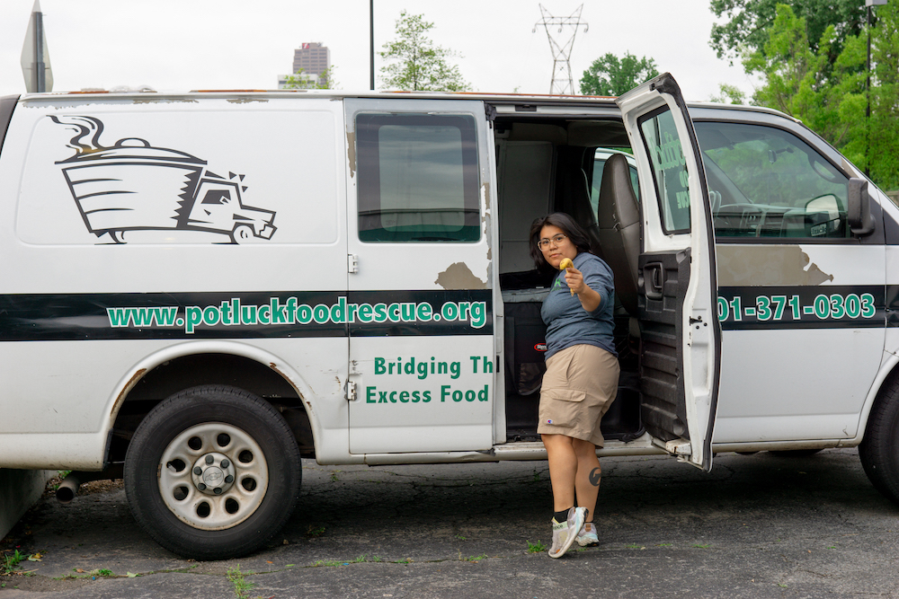 WeFightHunger A person with glasses and dark hair gives a thumbs up while standing in the open doorway of a Potluck Food Rescue van parked outdoors. The van has website and phone number details on the side.