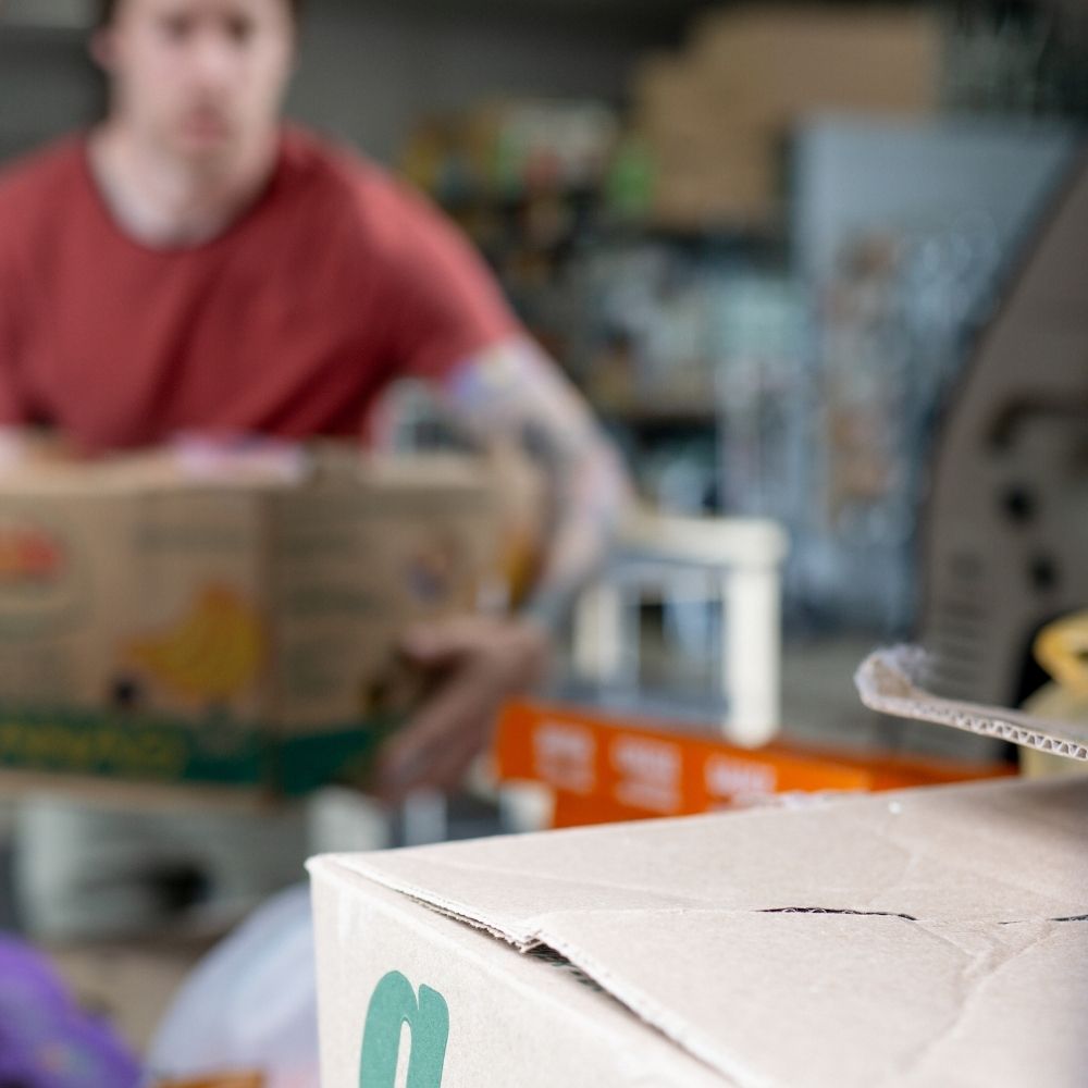 Who We Are Potluck A person in a red shirt carries a cardboard box in a room filled with other boxes and supplies, with the focus on a close-up cardboard box in the foreground.