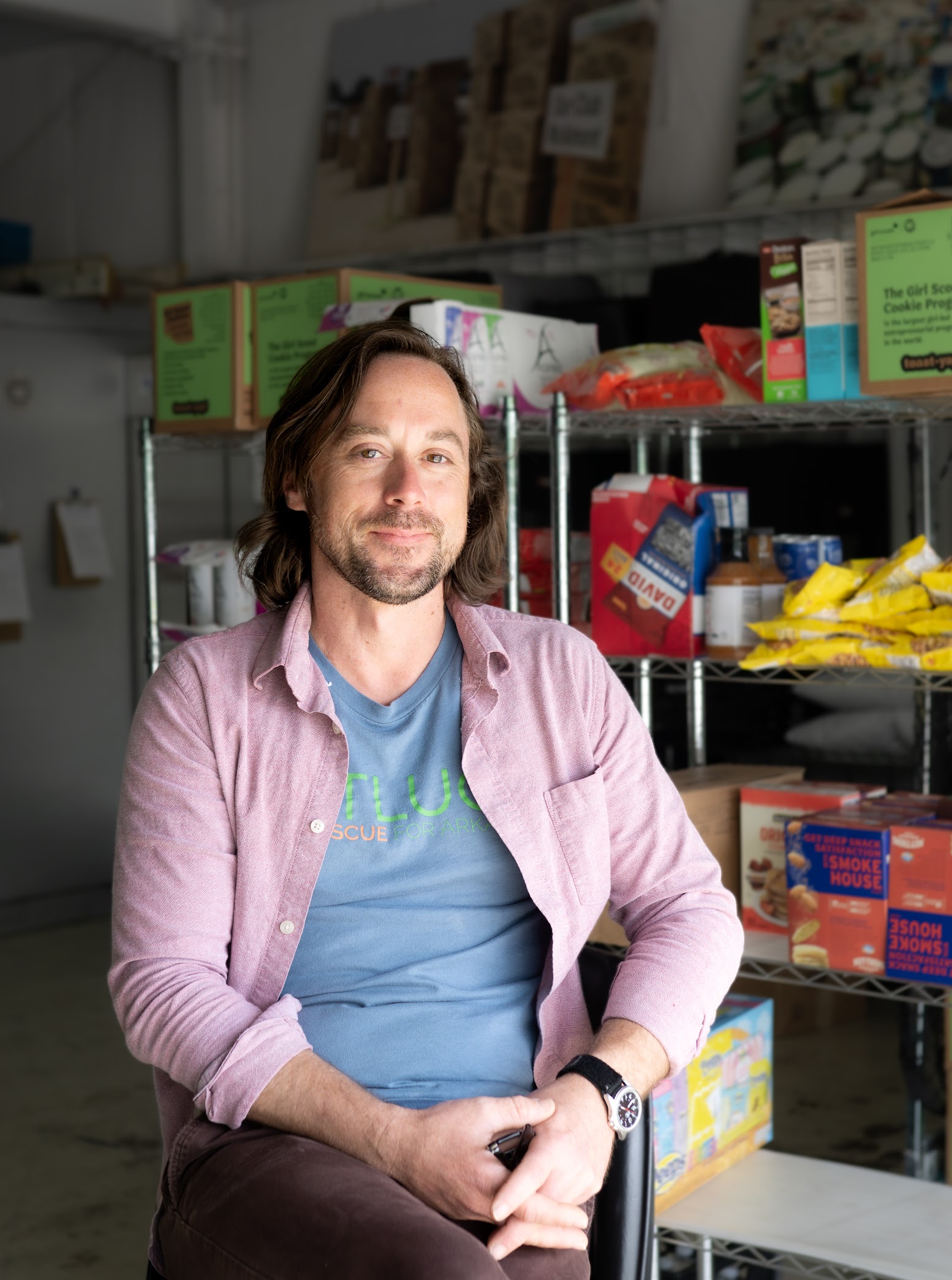 chrisheadshot1 A man with shoulder-length brown hair and a beard sits on a stool in front of shelves stocked with food and boxes, wearing a blue T-shirt, pink button-up shirt, and a watch, smiling at the camera.