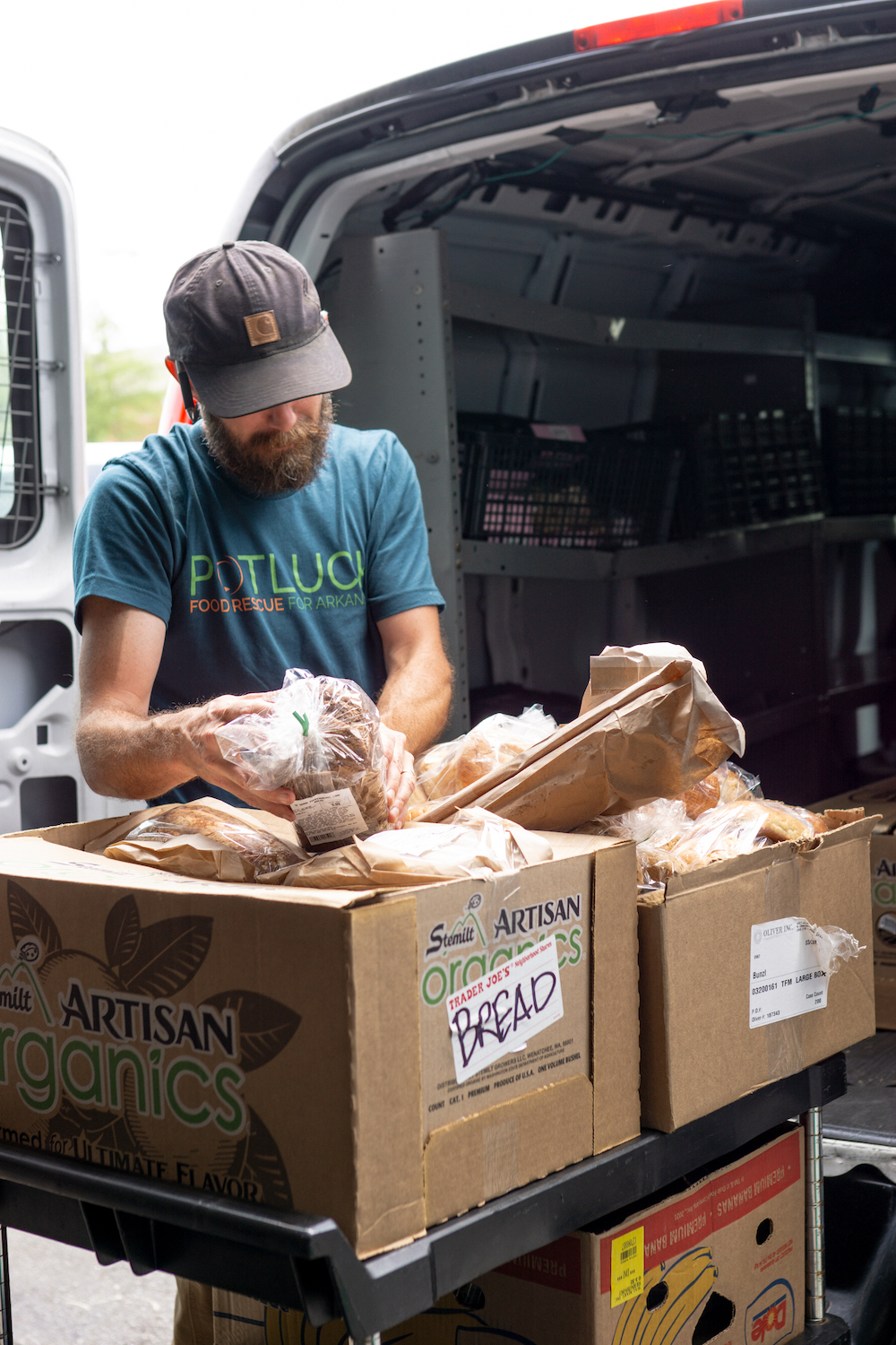 gettoknowus A man wearing a hat and a “POTLUCK” shirt sorts loaves of bread from cardboard boxes labeled “ARTISAN ORGANICS” near the open back of a delivery van.