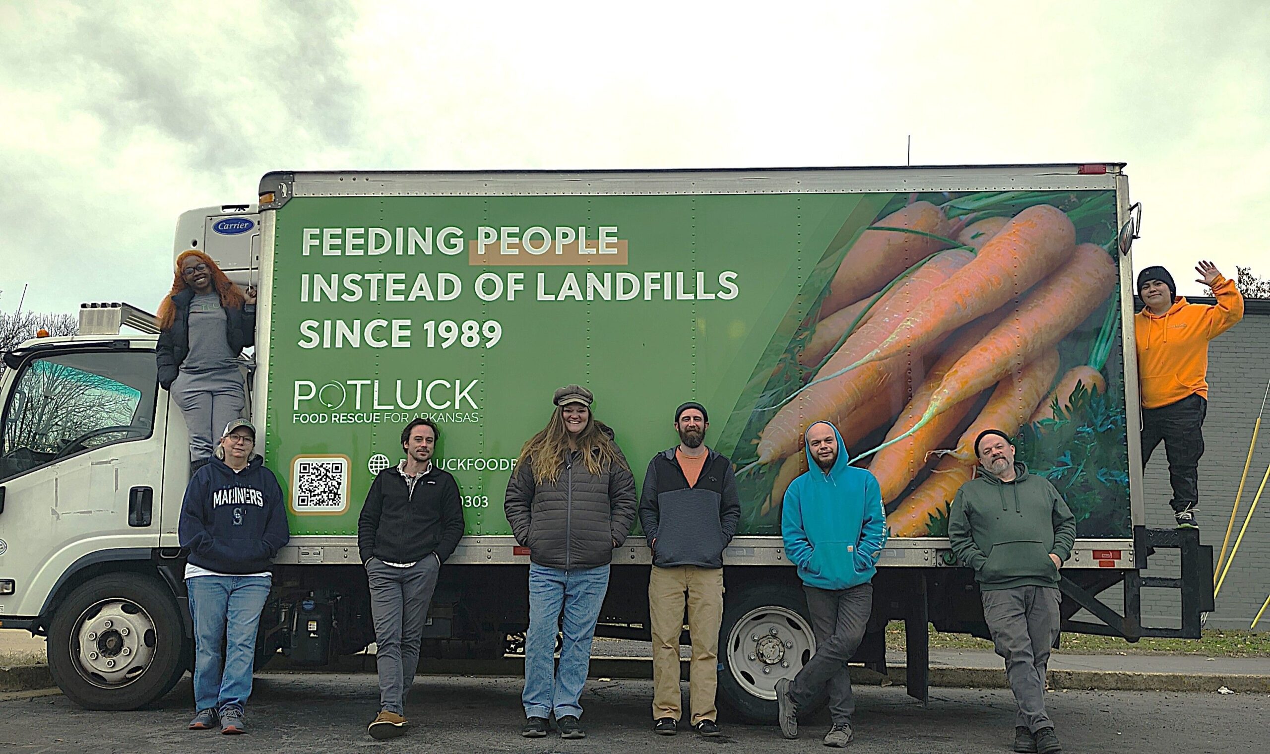 Eight people stand in front of a truck with a sign that reads, “Feeding people instead of landfills since 1989,” next to large carrots and the Potluck Food Rescue logo. Two people pose on the truck.