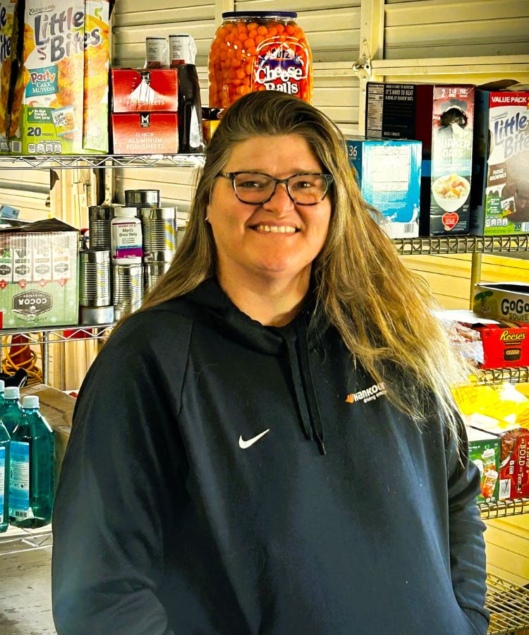 A smiling person with long hair and glasses wears a dark hoodie and stands in front of shelves stocked with snacks, drinks, and grocery items.