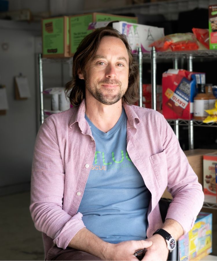A man with shoulder-length brown hair and a beard, wearing a light pink shirt over a blue t-shirt, sits and smiles in front of metal shelves stocked with food items in a storeroom.