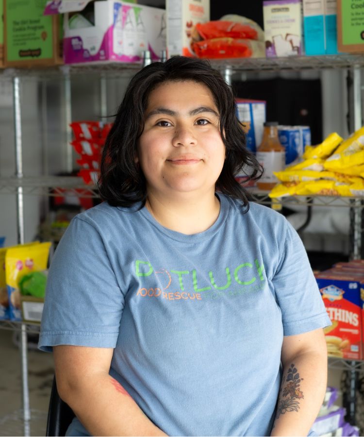 A person with dark hair wearing a blue “Potluck Food Rescue” shirt sits and smiles in front of shelves stocked with various packaged food items.