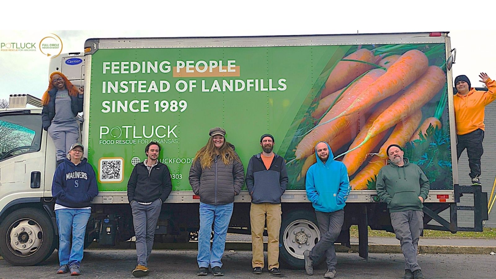Seven people pose in front of a truck decorated with large carrots and the text, Feeding People Instead of Landfills Since 1989. Potluck Food Rescue. Some stand, while two sit on the truck, all smiling outdoors.