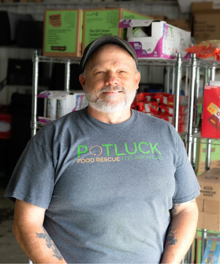 A smiling man with a gray beard stands in front of metal shelves holding boxes. He wears a cap and a Potluck Food Rescue for Arkansas t-shirt. The background appears to be a storage area or warehouse.
