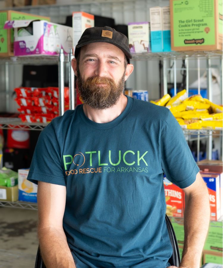 A bearded man in a POTLUCK Food Rescue for Arkansas shirt and black cap sits smiling in front of shelves stocked with food boxes and snacks in a storage area.