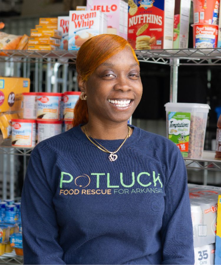 A smiling woman stands in front of shelves stocked with food. She wears a navy “Potluck: Food Rescue for Arkansas” shirt and gold jewelry. Various food items, including Dream Whip and Nut-Thins, are visible behind her.